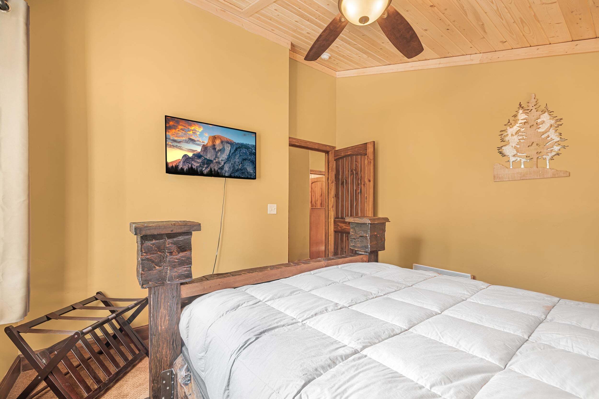 A bedroom with a bed, a wooden headboard, a mounted TV, a wooden tree decoration, and a mountain painting on the wall.
