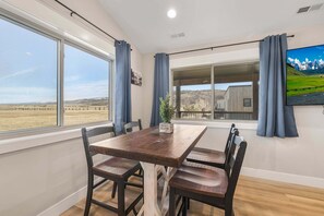 A dining area with a wooden table and chairs, large windows with curtains, and a view of a rural landscape. - Victor Traverse (Victor)