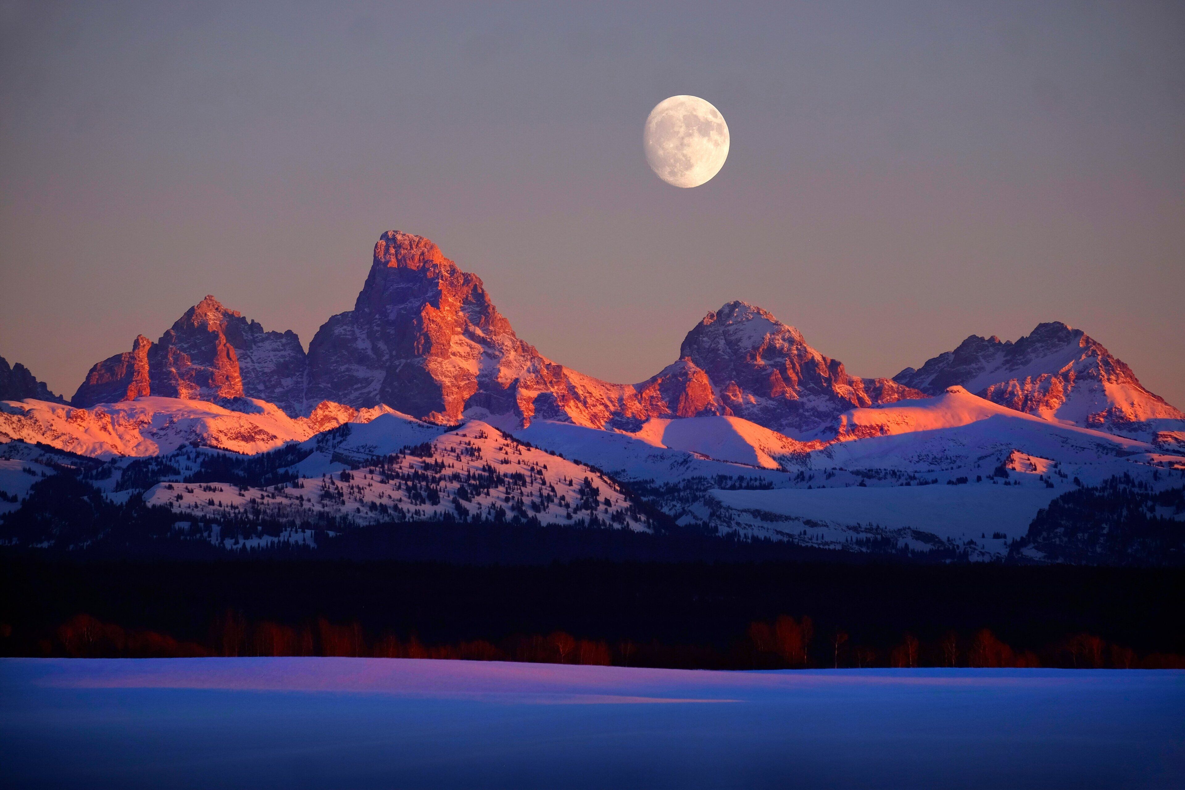 Paisaje montañoso con picos nevados y una luna llena en el cielo.