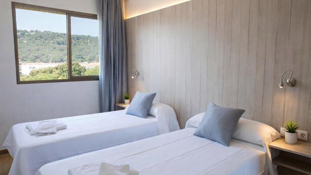 Une chambre d’hôtel avec deux lits, une fenêtre donnant sur un espace verdoyant, une table de chevet avec une lampe et une prise électrique fixée au mur.