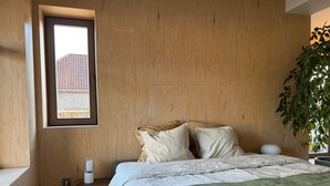 A bedroom with a bed, pillows, a bedside table, a lamp, a window, and a plant.