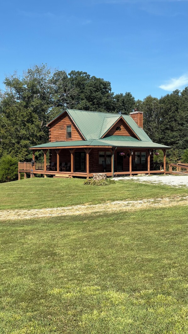 A two-story log cabin with a green metal roof, a covered porch with outdoor seating, and a grassy yard.
