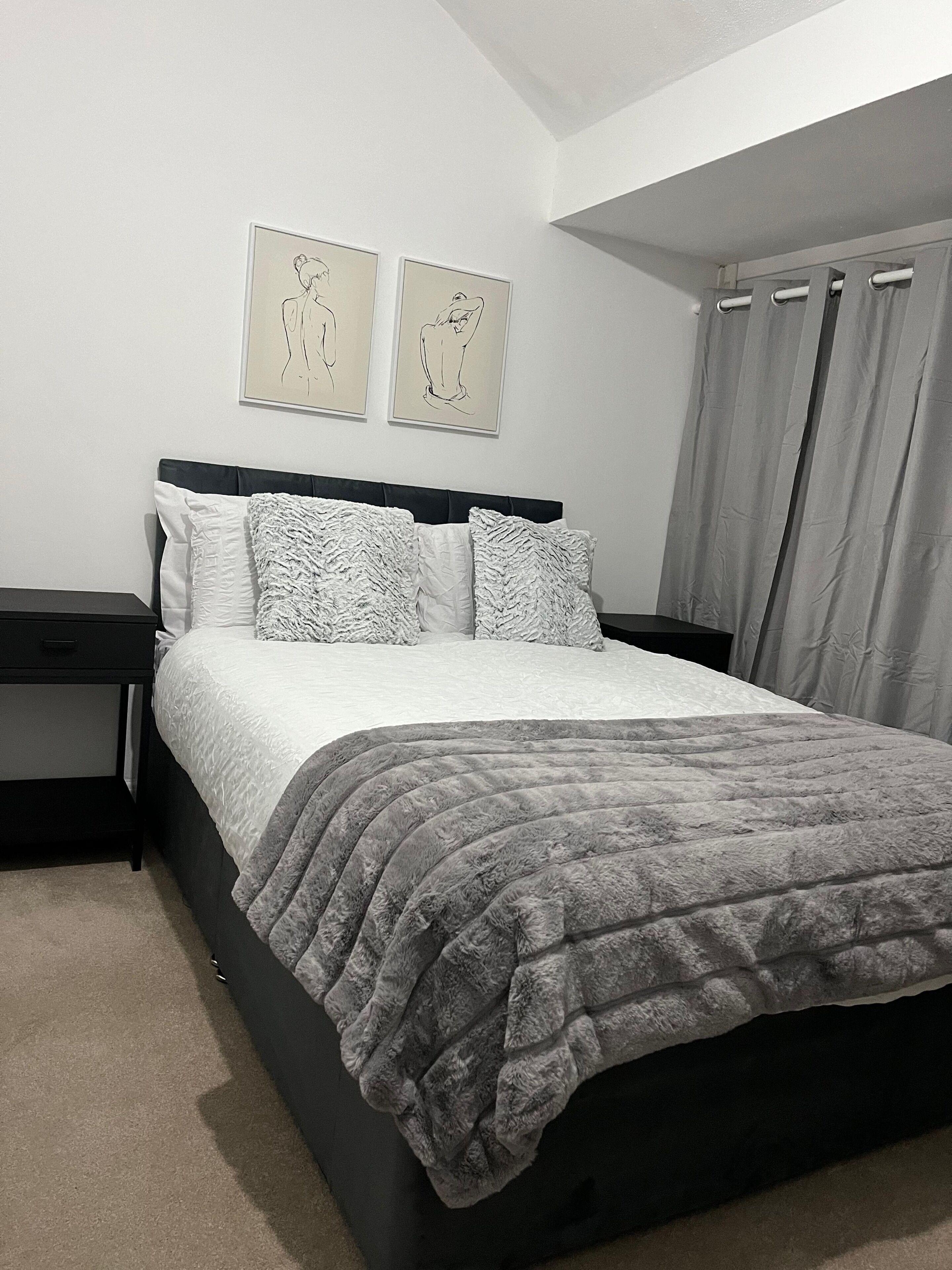 A bedroom with a bed, two nightstands, a headboard, a window with curtains, and two wall-mounted artworks.