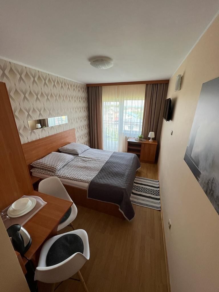 Ein kleines Hotelzimmer mit Bett, Schreibtisch mit Stuhl, Nachttisch mit Lampe und einem Fenster mit Vorhängen.