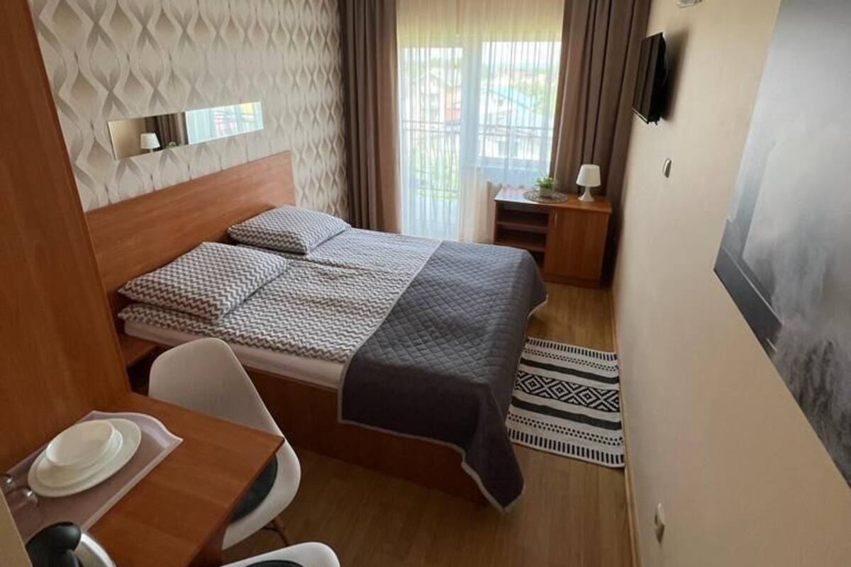 A compact hotel room with a bed, a desk with a chair, a small table with a lamp, and a window with curtains.