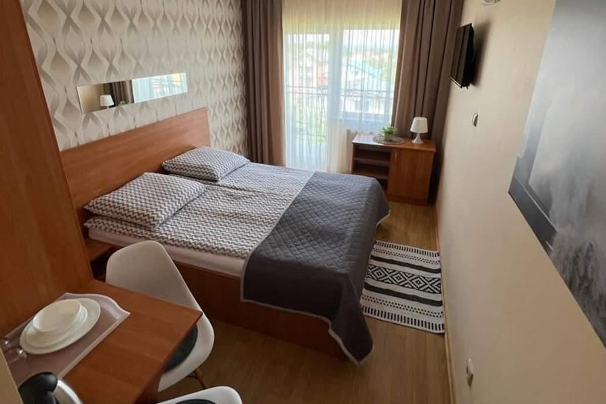 A compact hotel room with a bed, a desk with a chair, a small table with a lamp, and a window with curtains.