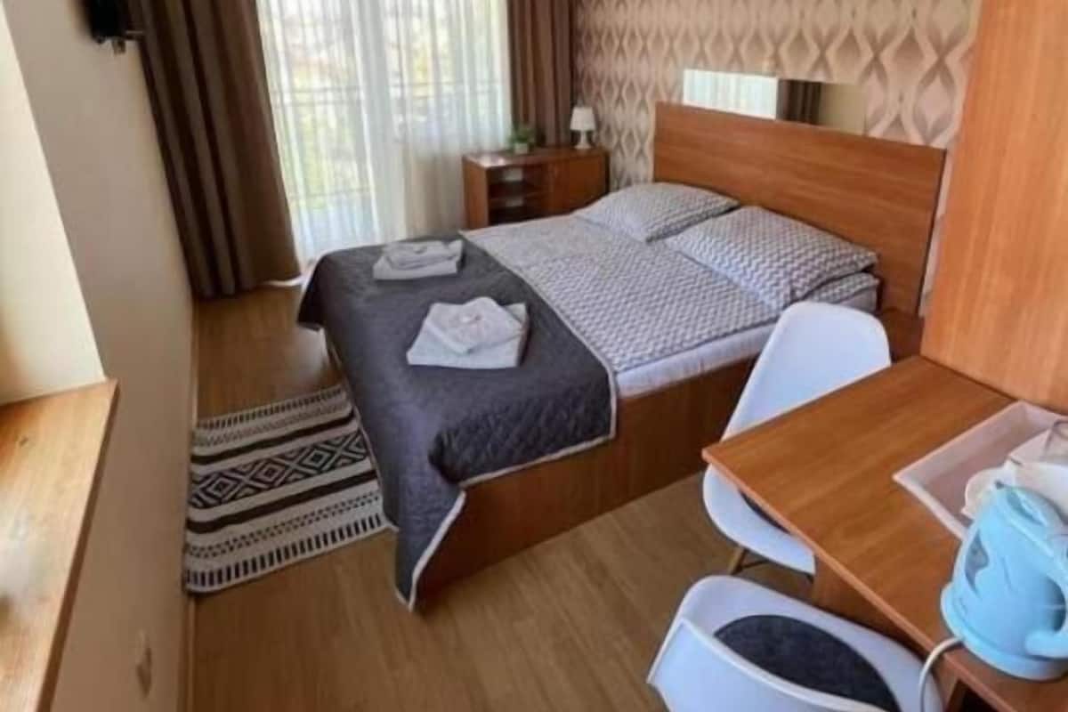 A compact hotel room with a bed, bedside table, chair, desk, and a small table with a kettle.