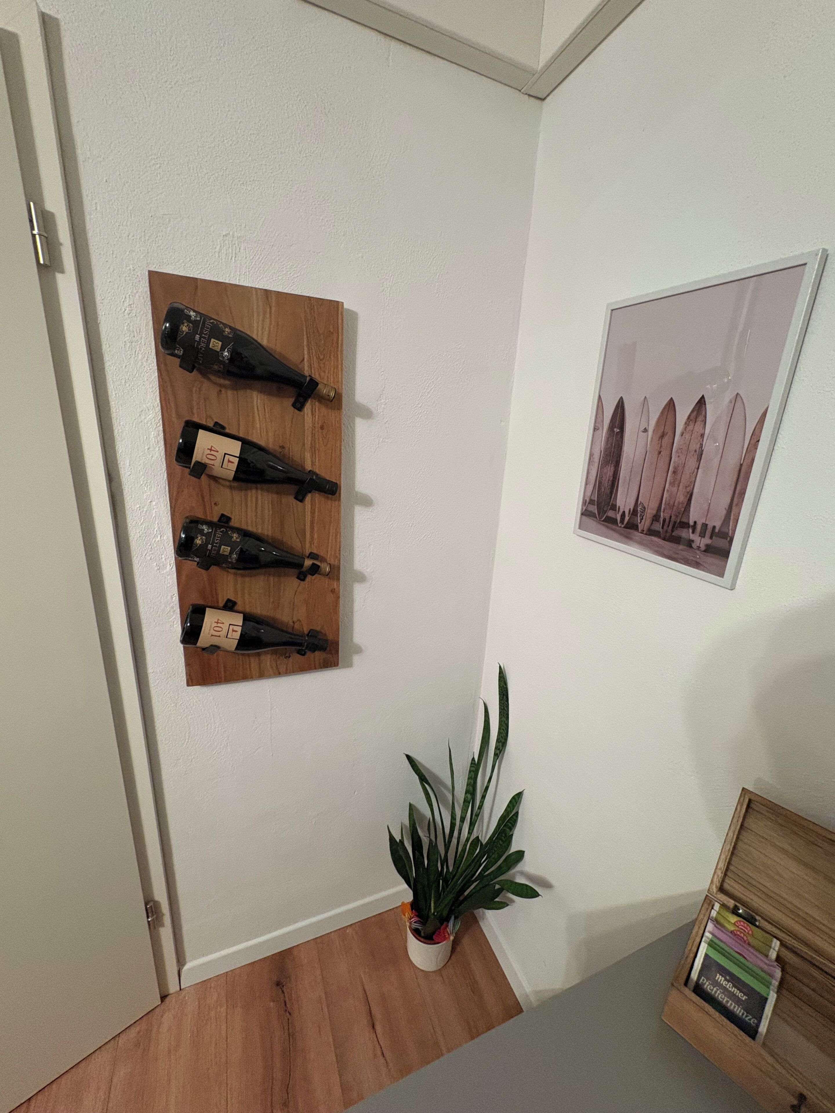 Ein Zimmer mit einem an der Wand montierten Weinregal aus Holz, das vier Flaschen beherbergt, einem gerahmten Bild, einer Topfpflanze und einem Holzschrank mit Büchern.