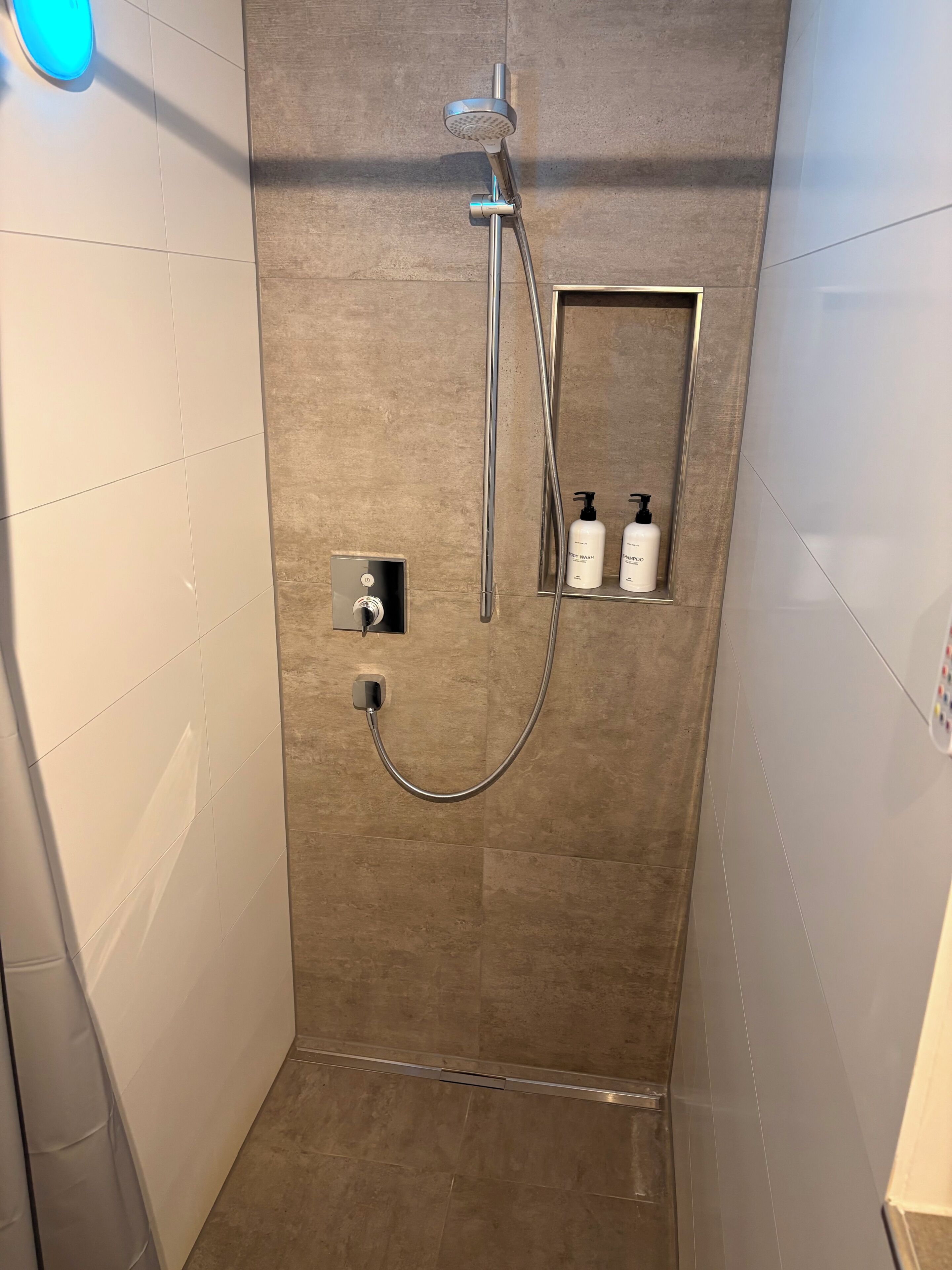Eine Dusche mit Duschbrause, Wandarmatur und Bedienfeld. Auf einem Regal stehen zwei Flaschen Shampoo und Conditioner.