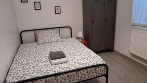 A bed with a patterned bedspread, a pillow, a folded blanket, and a bedside lamp. There are three framed pictures on the wall, a wardrobe, and a radiator. - First floor room in a town house. (La Riche)