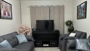 A living room with two grey sofas, a flat-screen TV on a black stand, a ceiling fan, and two wall-mounted paintings.