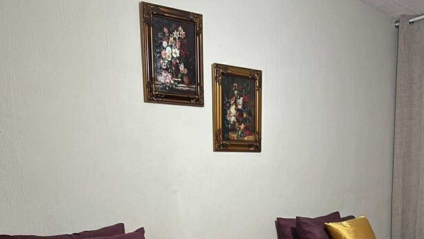 A hotel room with two beds, each with red bedding and multiple pillows. There are two framed floral paintings on the wall.