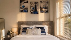 Ein ordentlich bezogenes Bett mit Holz Kopfteil, ein Nachttisch mit Lampe und Wandbild mit Weingläsern und Rosen.
