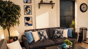 Ein Wohnzimmer mit einem dunklen Sofa, gemusterten Kissen, einem Couchtisch mit Pflanze, einer Lampe und Kunstwerken an der Wand.