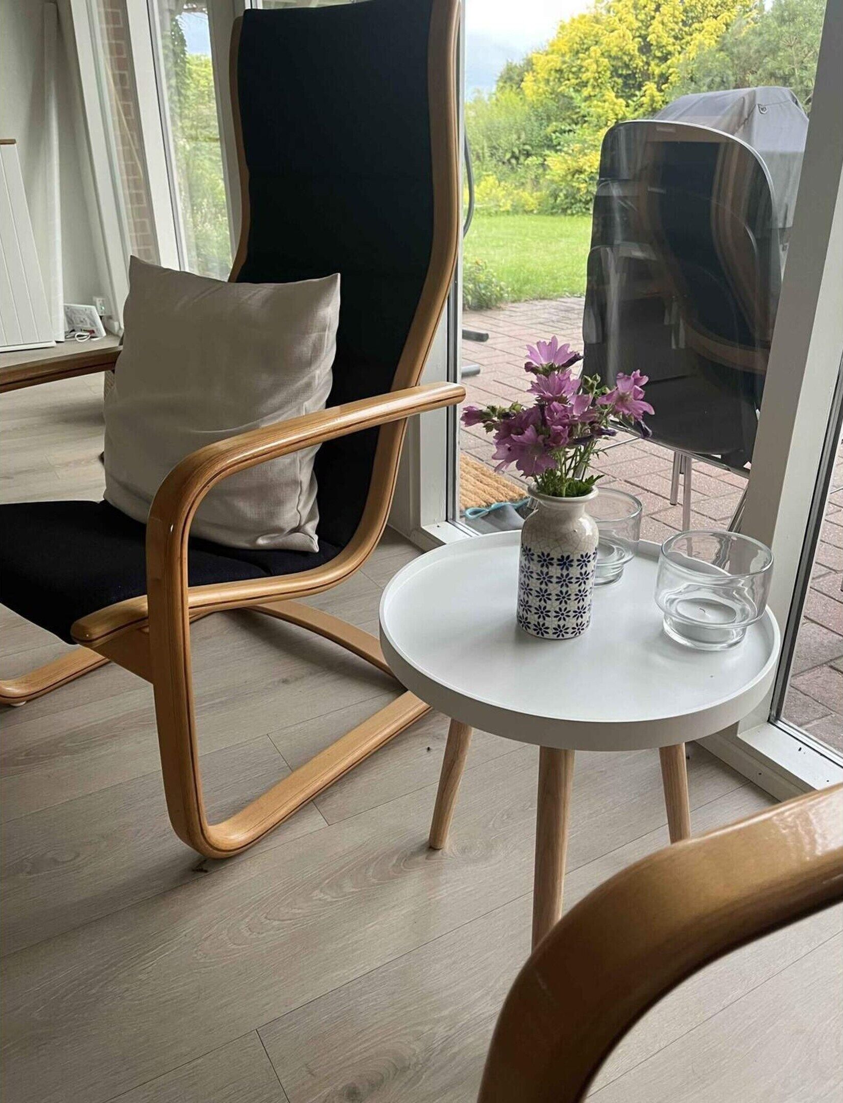 A modern room with a wooden chair, a small round table with a vase and glasses, and a view of the outdoors through a glass door.