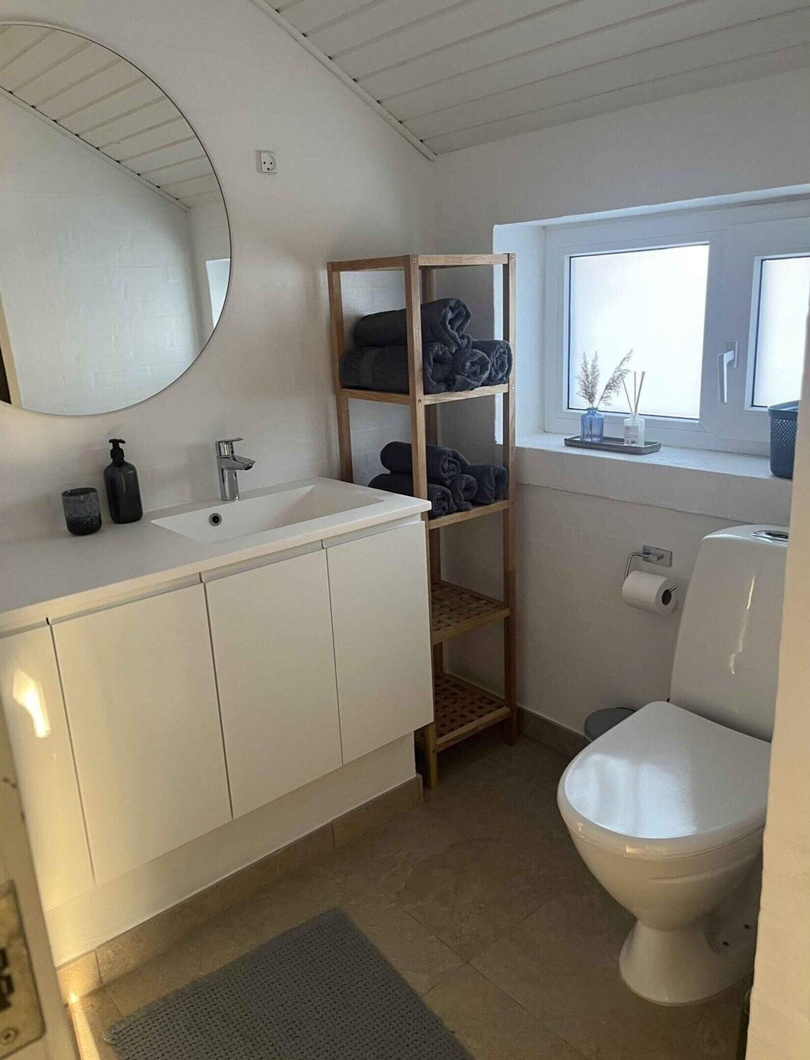 Een badkamer met een wit wastafel, spiegel en toilet. Er is een houten plank met handdoeken en een raam boven het toilet.