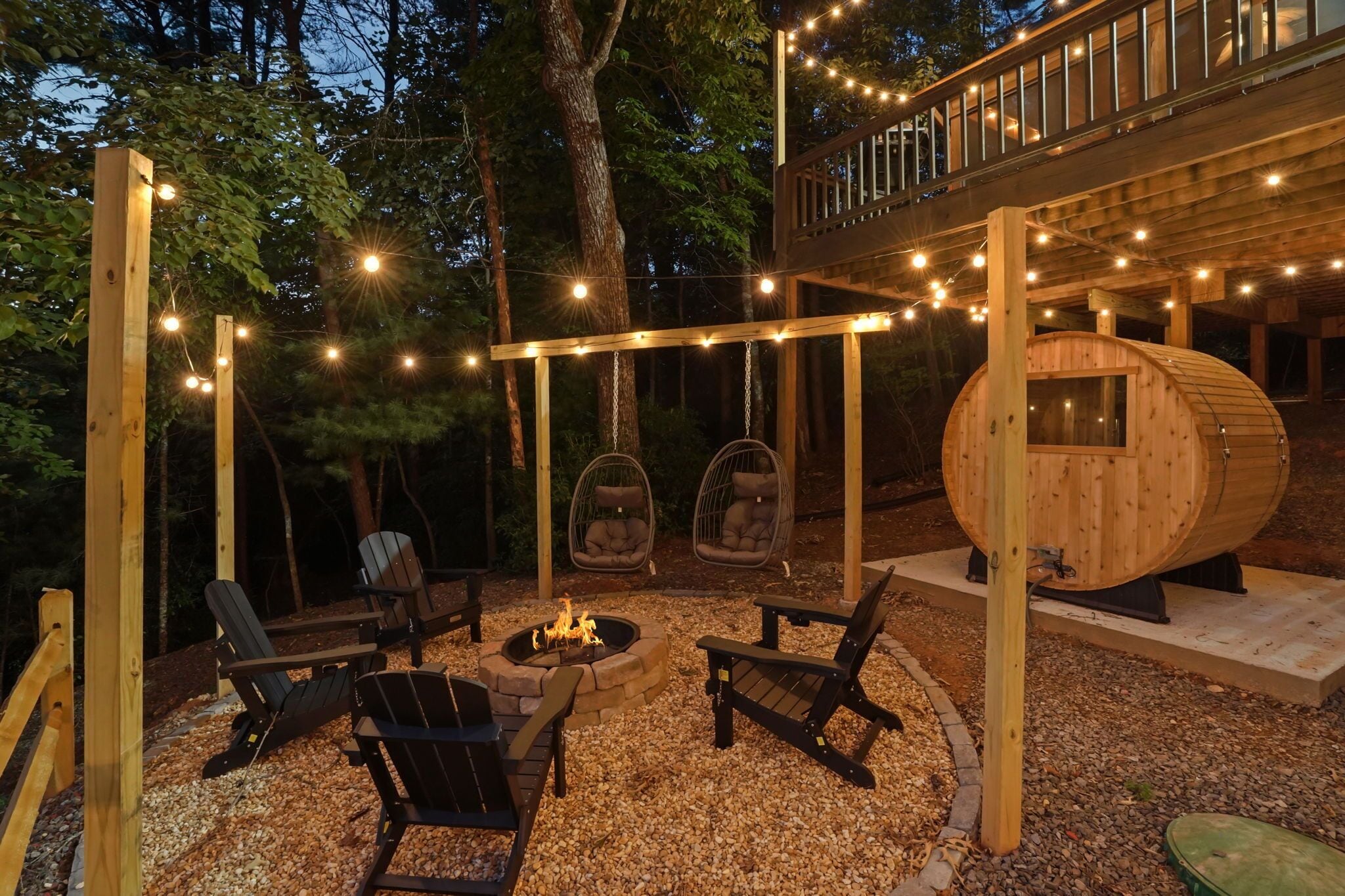 A wooden deck with string lights, a fire pit, Adirondack chairs, and a wooden sauna.