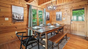 Eine Holzhütte mit Esstisch für vier Personen, einer Kristallleuchtre und zwei mit Vorhängen versehenen Fenstern.