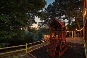 Eine hölzerne Spielkonstruktion mit Rutsche und Kletterwand, umgeben von Bäumen und einer Terrasse mit Lichterketten.