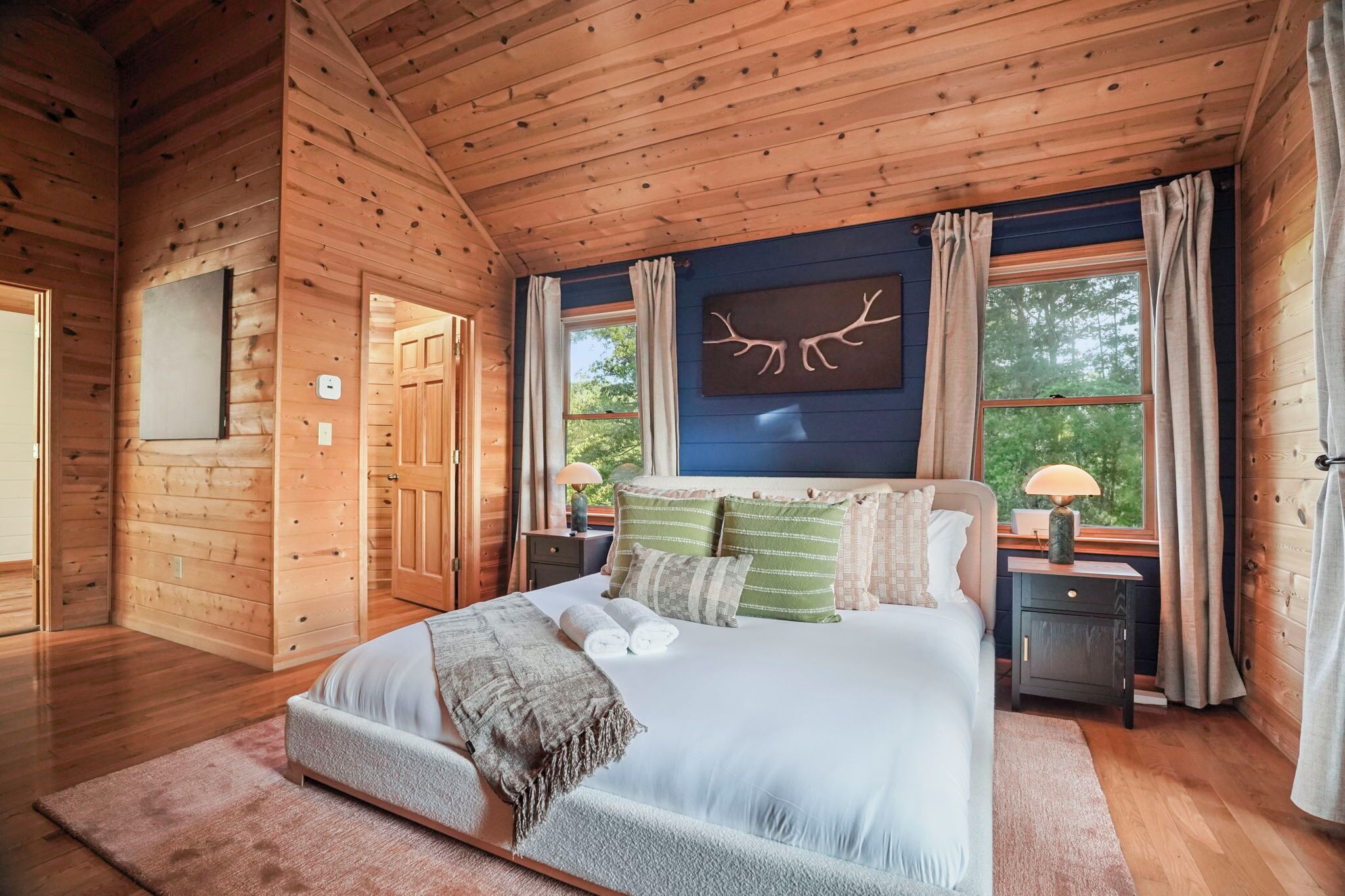 A wooden cabin bedroom with a bed, nightstands, a window with curtains, and a wall-mounted artwork.