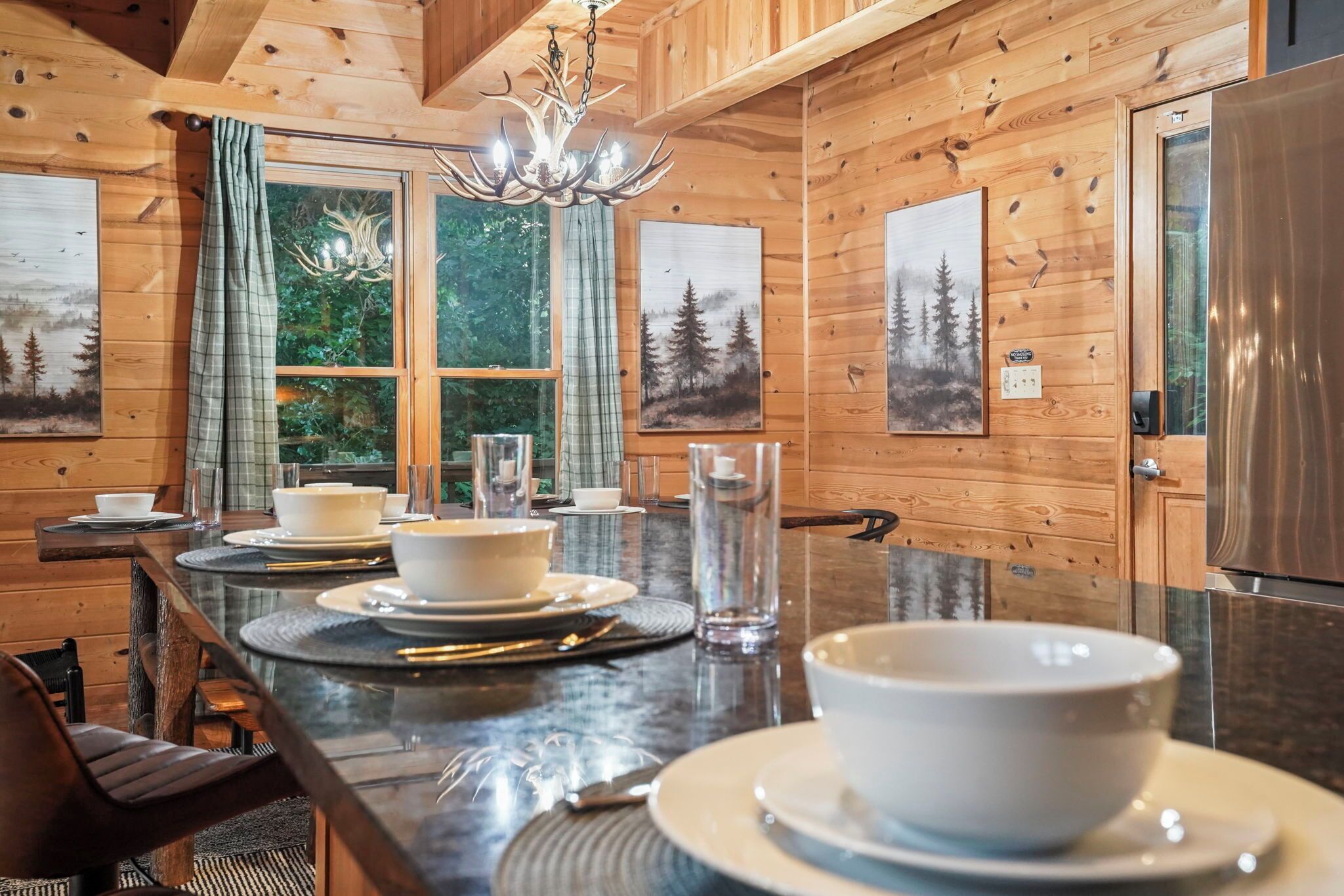 A wooden interior with a dining table set for four, surrounded by chairs, and a large window with curtains.