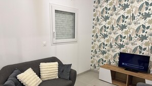 Ein Wohnzimmer mit grauem Sofa, weißen und grauen Kissen, einem Holztisch, einem Fernseher auf einem Holzmöbel und einem Fenster mit weißem Rollo.