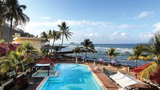 Piscina de un complejo turístico con vista al océano, rodeada de palmeras y una playa.