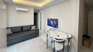 Una sala de estar moderna con un sofá, una mesa de comedor con sillas, un televisor montado y luces empotradas en el techo.