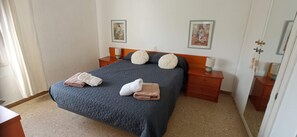 Ein ordentlich bezogenes Bett mit zwei Kissen, ein Nachttisch mit Lampe und zwei an der Wand hängende Bilder.