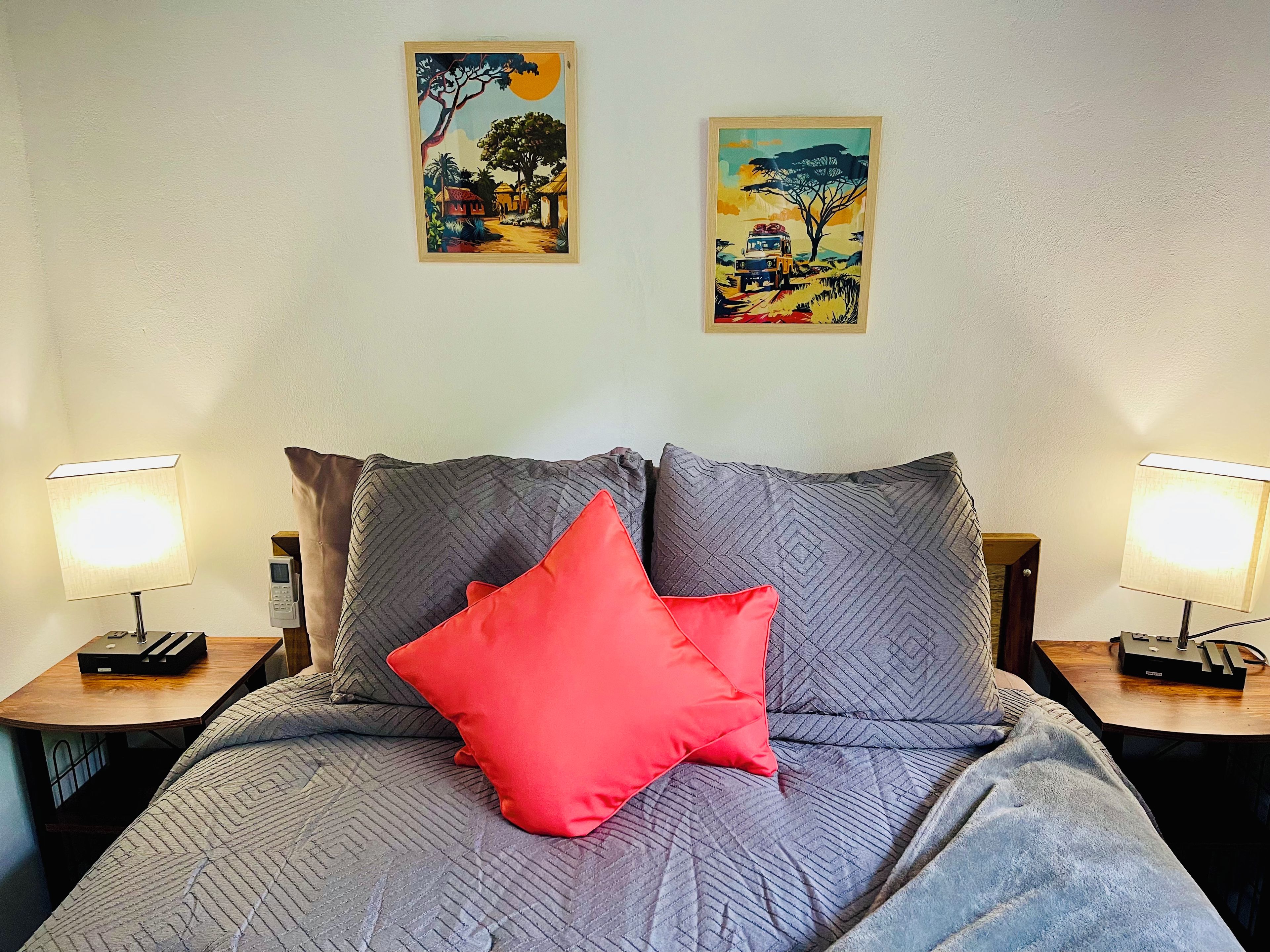 Ein Bett mit grauer Bettwäsche und einem roten Kopfkissen, flankiert von zwei Nachttischen mit Lampen, vor einer Wand mit zwei gerahmten Kunstwerken.