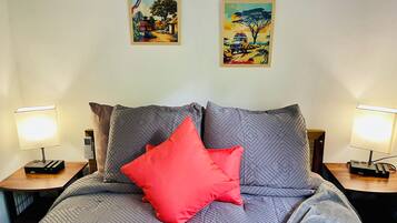 Un lit avec une literie grise et un oreiller rouge, flanqué de deux tables de chevet avec des lampes, le tout adossé à un mur où sont accrochées deux œuvres d’art encadrées.