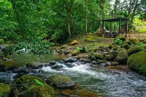 Un ambiente naturale con un ruscello, rocce e un gazebo in legno con posti a sedere.