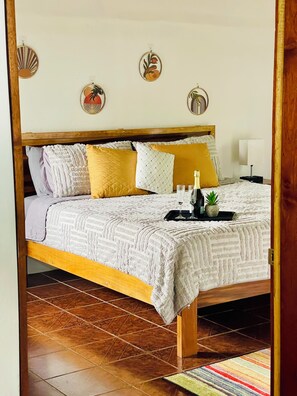 Un lit avec un cadre en bois, plusieurs oreillers, une table de chevet avec une lampe et des œuvres d’art décoratives accrochées au mur.