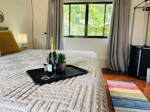 A neatly made bed with a bottle of champagne and two glasses on a black tray, a small potted plant, and a colorful striped rug on the floor. - New! Oasis-On River with King bed & Double bed, Air Conditioner, Hot water,  (La Fortuna)