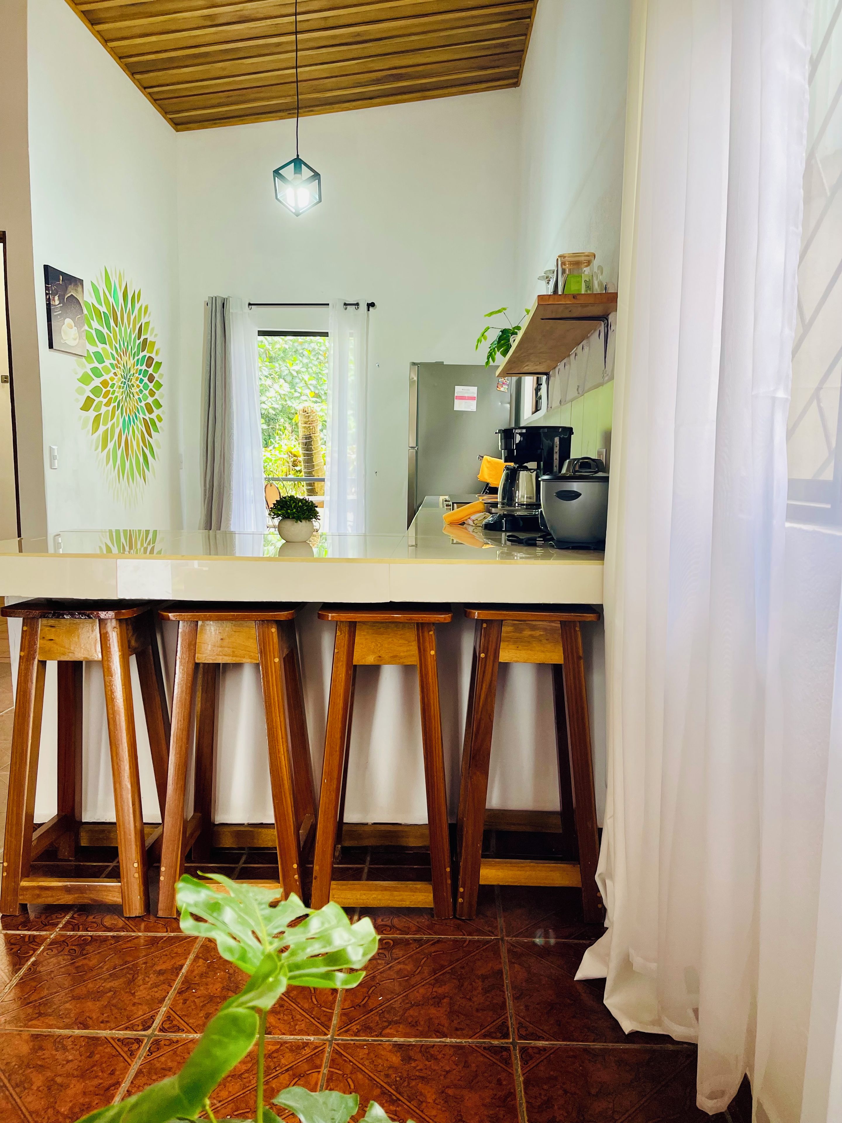Cucina con soffitto in legno, un piano di lavoro bianco, sgabelli di legno, un lampadario a sospensione e vista su piante.