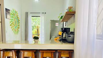 Cucina con soffitto in legno, un piano di lavoro bianco, sgabelli di legno, un lampadario a sospensione e vista su piante.