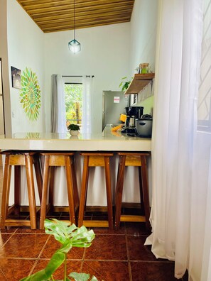 Cucina con soffitto in legno, un piano di lavoro bianco, sgabelli di legno, un lampadario a sospensione e vista su piante.