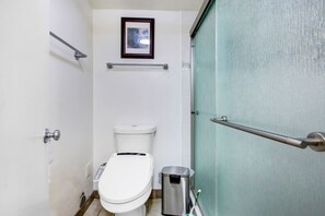 Een kleine badkamer met toilet, een prullenbak, een lijst met foto en een metalen handgreep.