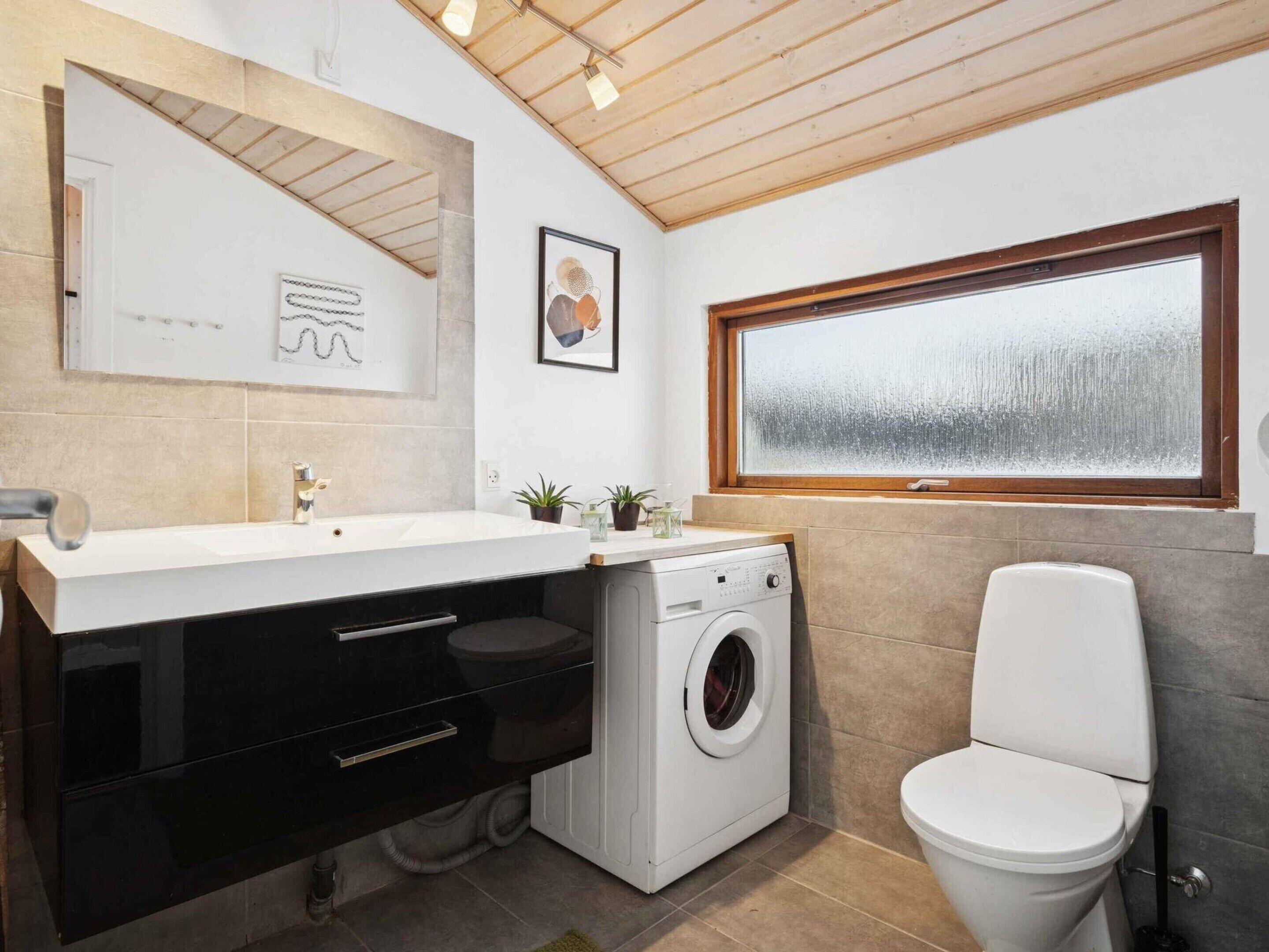 A modern bathroom with a sink, mirror, washing machine, toilet, and a window.