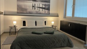 Un dormitorio con cama, dos mesitas de noche, un radiador, una ventana con persianas y un cuadro grande con un paisaje en la pared.