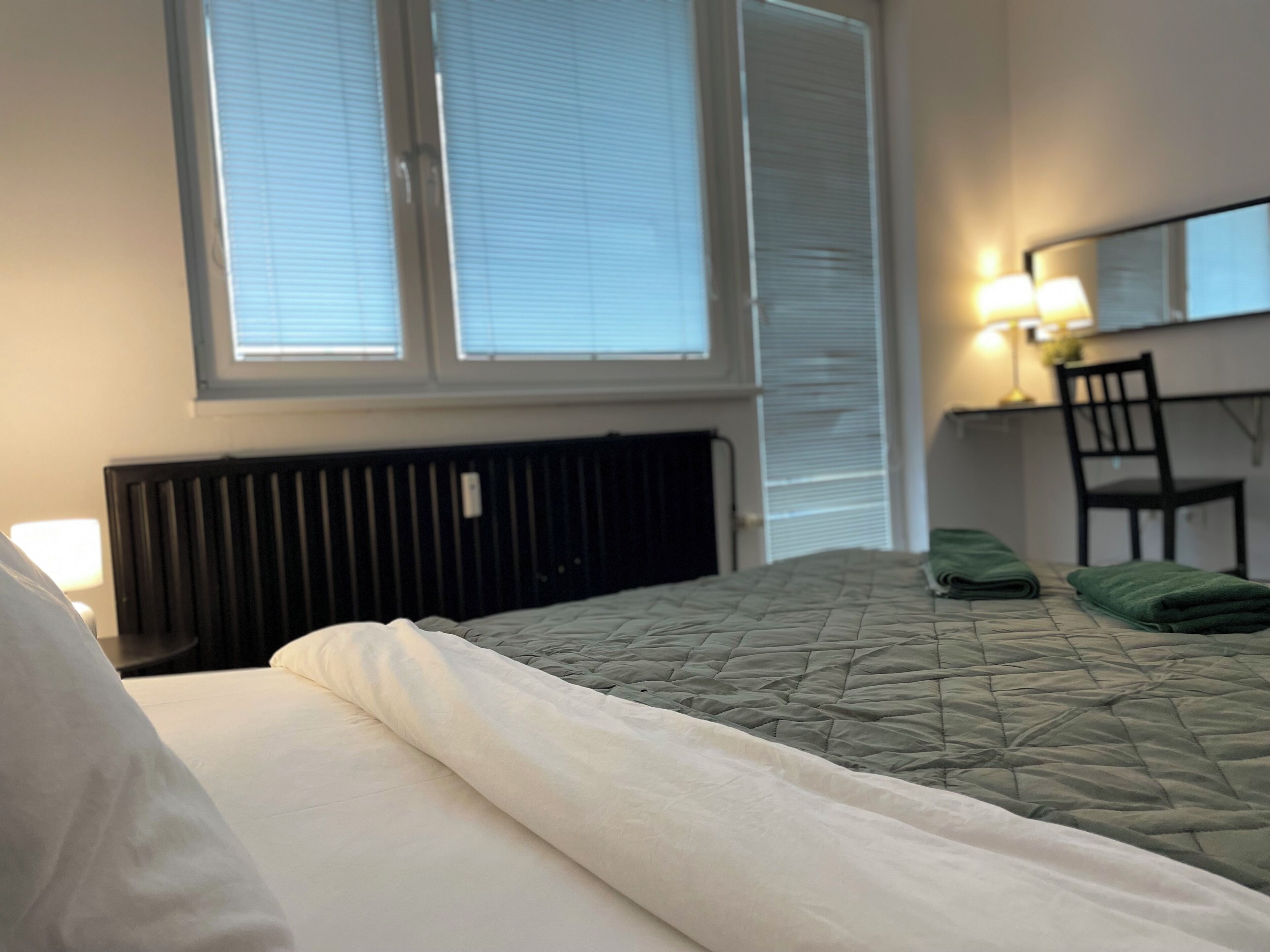 A hotel room with a bed, a chair, a desk with a lamp, and a window with blinds.