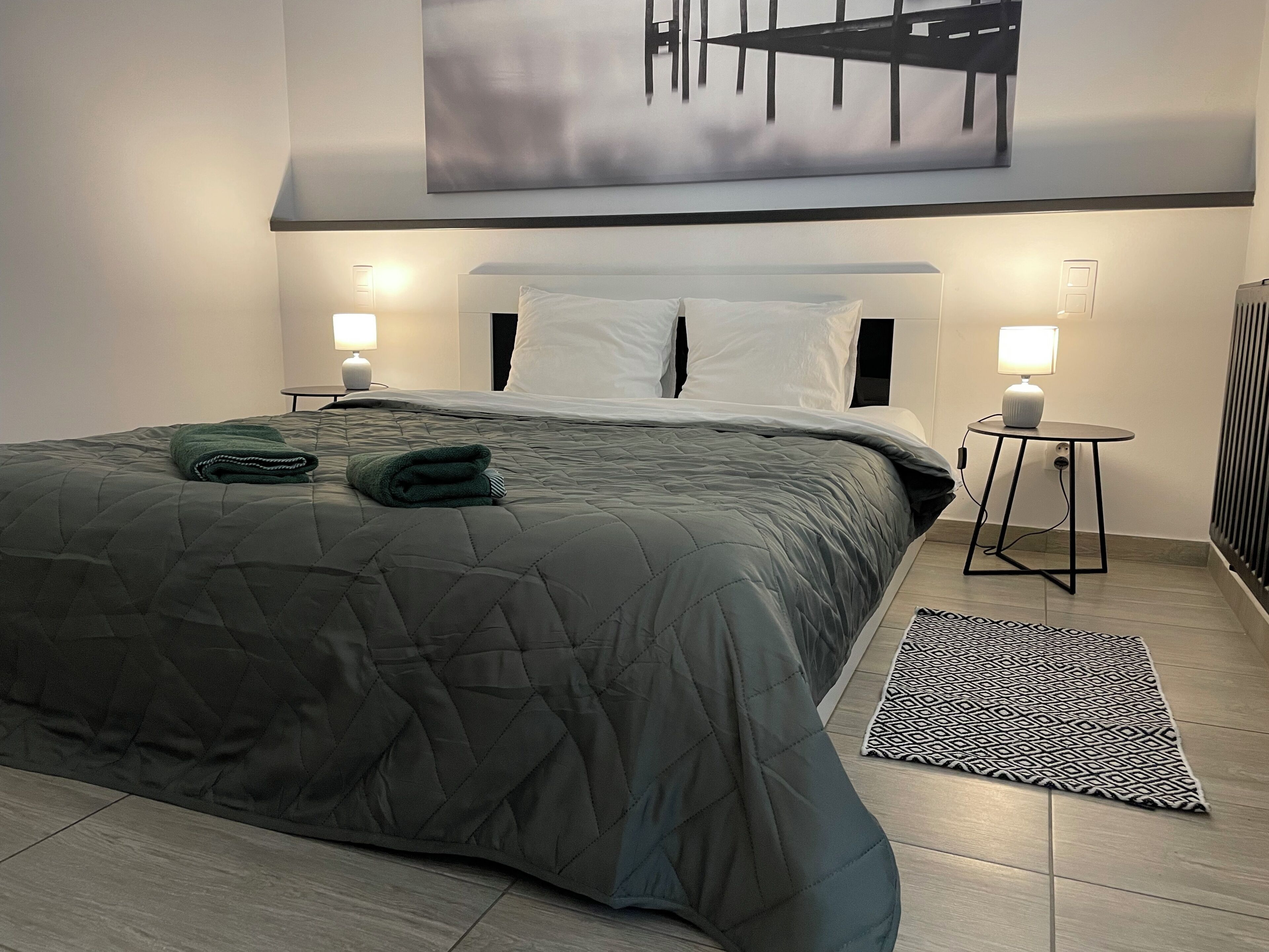 Een netjes opgemaakt bed met een grijs geborduurd dekbed, twee bedlampen, een zwart-wit gestreept tapijt en een groot abstract schilderij aan de muur.