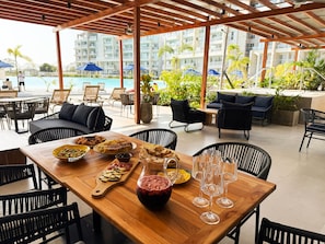 A wooden table with food, wine, and glasses, surrounded by chairs and umbrellas, overlooking a pool and buildings. - Stunning Lagoon View Condo w/ Beach, Pool & Jacuzzi (Cartagena)