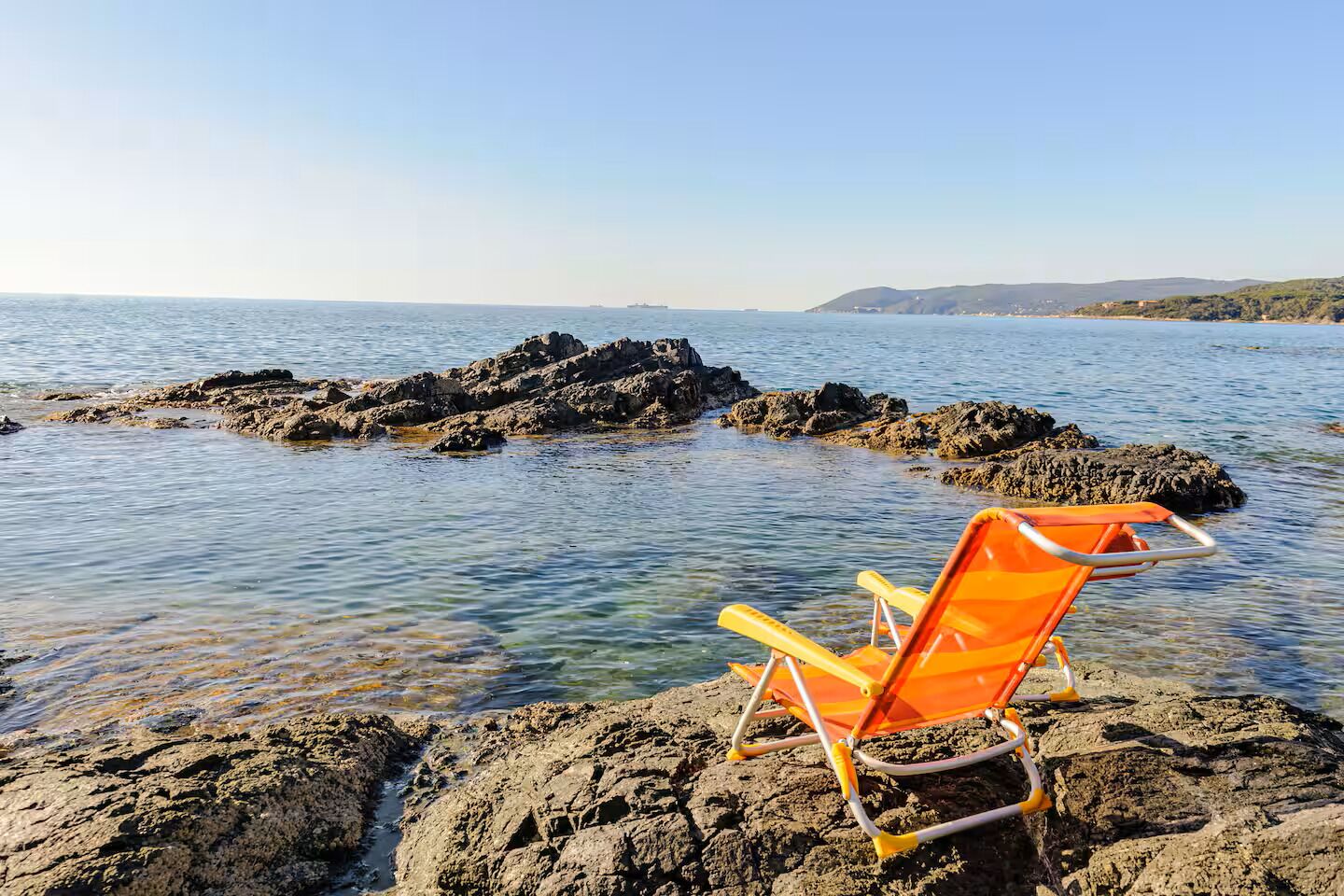 A chair on rocky terrain near calm water with distant land.