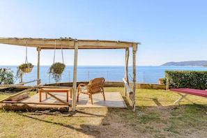 Eine hölzerne Pergola mit einem Korbstuhl und einem Tisch mit Blick auf die Küste.