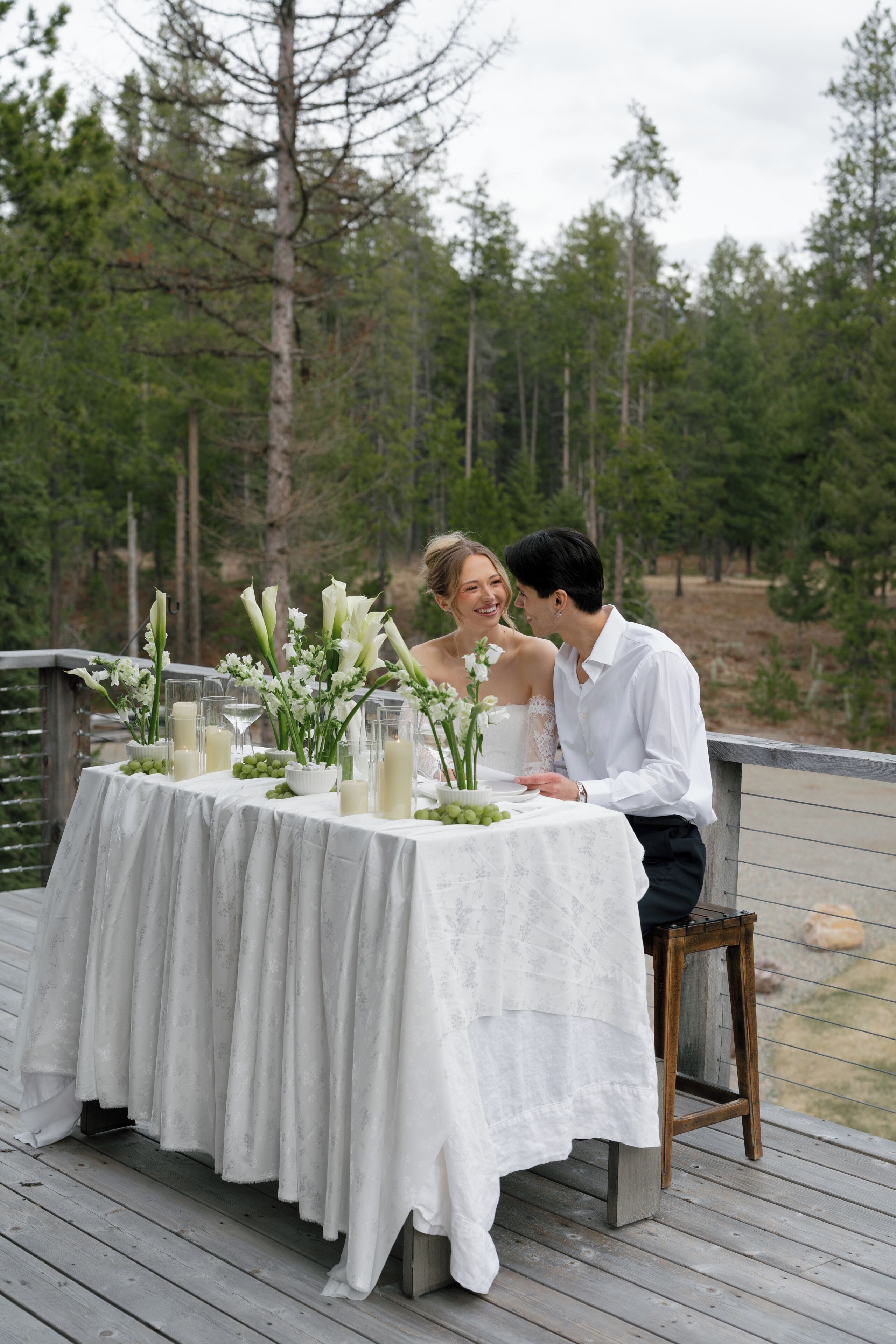 A couple seated at a table with a white tablecloth, surrounded by greenery and trees.