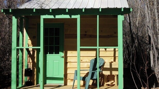 A wooden cabin with a green door, a window, and a small porch with a chair.