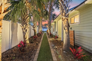 A pathway flanked by palm trees and a white fence leading to a house with a window. - 3BR Pool & Hot Tub Retreat | Good Morning Rentals (Jacksonville Beach)