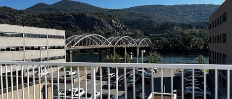 Vista desde un balcón con barandilla, que da sobre un estacionamiento y un puente sobre el agua, con montañas al fondo.