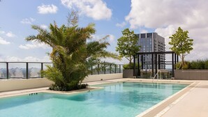 En pool på taket med fri utsikt över stadens silhuett, omgiven av palmer och en modern byggnad.